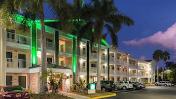 Un hotel de varios pisos con estacionamiento, palmeras y fachada iluminada en verde.