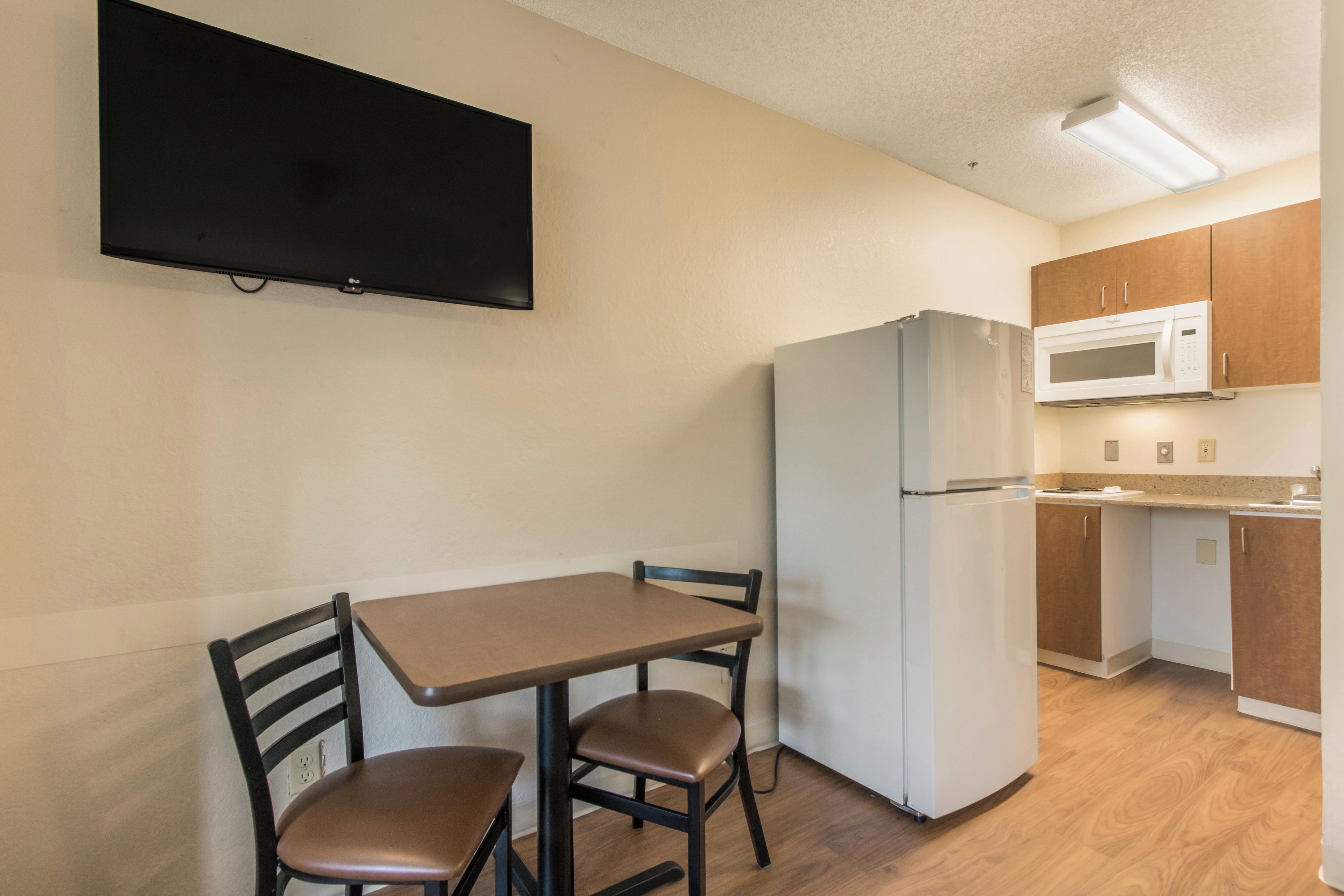 A compact kitchen with a dining table, chairs, a refrigerator, a microwave, and a flat-screen TV mounted on the wall.