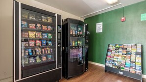 Máquina expendedora con aperitivos, dispensador de agua y una pequeña selección de libros de viaje, junto a una pared verde con una cámara de seguridad.