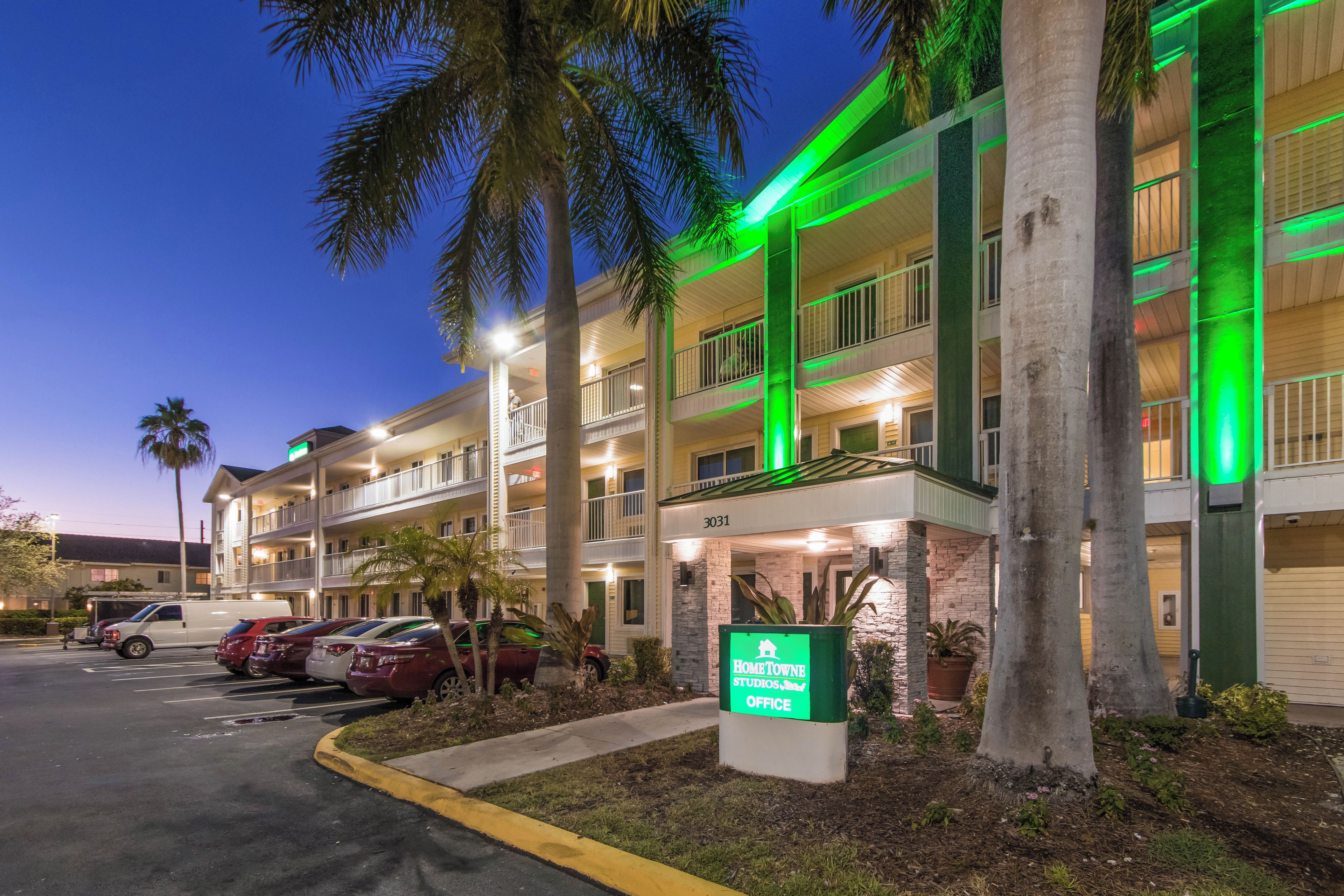 A multi-story hotel with a parking lot, palm trees, and a sign for the Hometown Studios Office.
