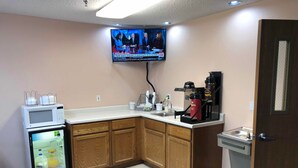 Cocina compacta con microondas, refrigerador, cafetera y televisor montado en la pared mostrando noticias.