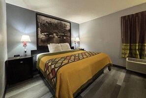 A hotel room with a bed, two bedside tables, a nightstand, a wall-mounted lamp, a large framed picture, and an air conditioning unit.