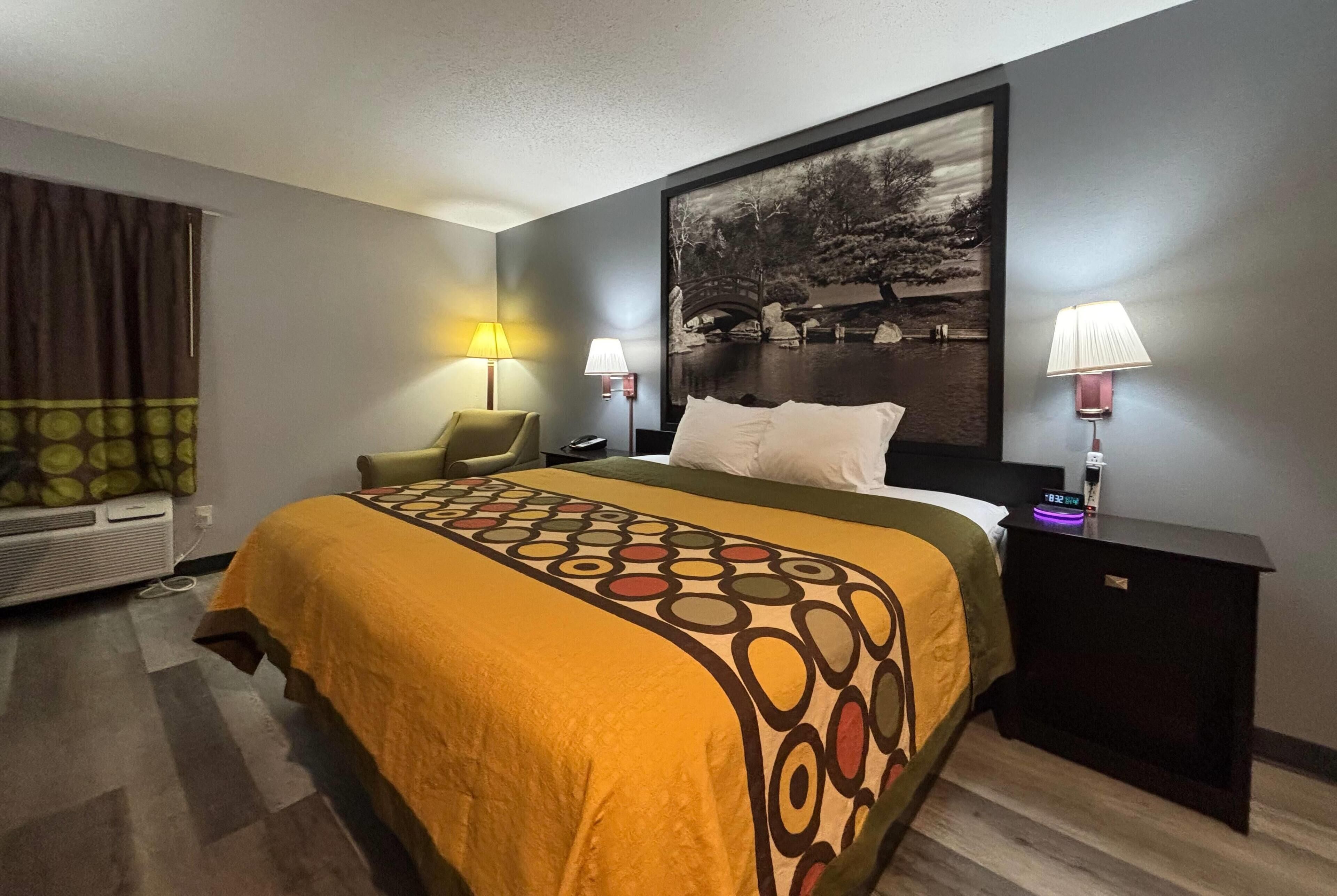 A hotel room with a bed, two bedside lamps, a chair, a wall-mounted heater, and a large framed picture.