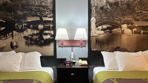Una habitación de hotel con dos camas, una mesita de noche con lámpara, teléfono y un mural en la pared que muestra un puente sobre el agua.