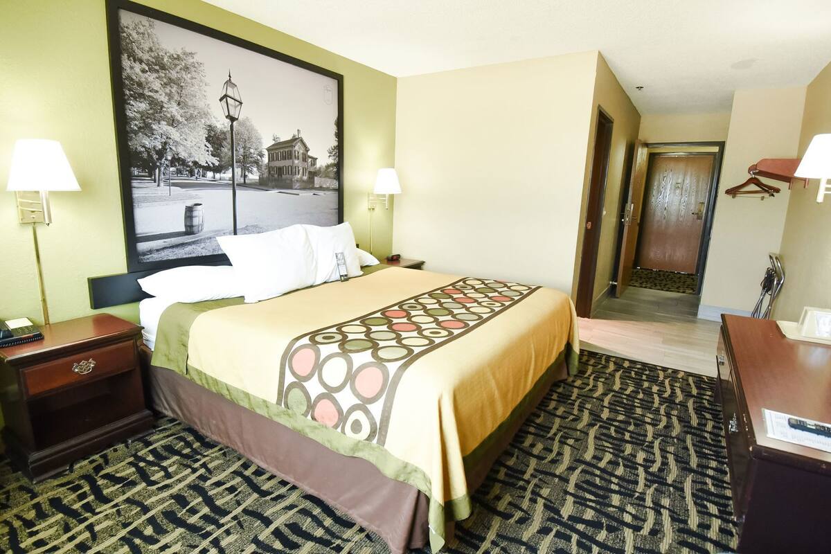 Ein Hotelzimmer mit Bett, zwei Nachttischen, einem Schreibtisch, einem Sessel, einem gemusterten Teppich, einem gerahmten Bild an der Wand und einer Tür zu einem anderen Raum.
