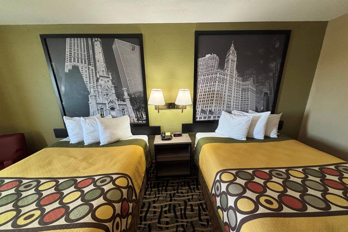 Ein Hotelzimmer mit zwei Betten, jeweils mit einem gemusterten Überwurf, einem Nachttisch mit Lampe sowie zwei gerahmten Bildern von Gebäuden an der Wand.