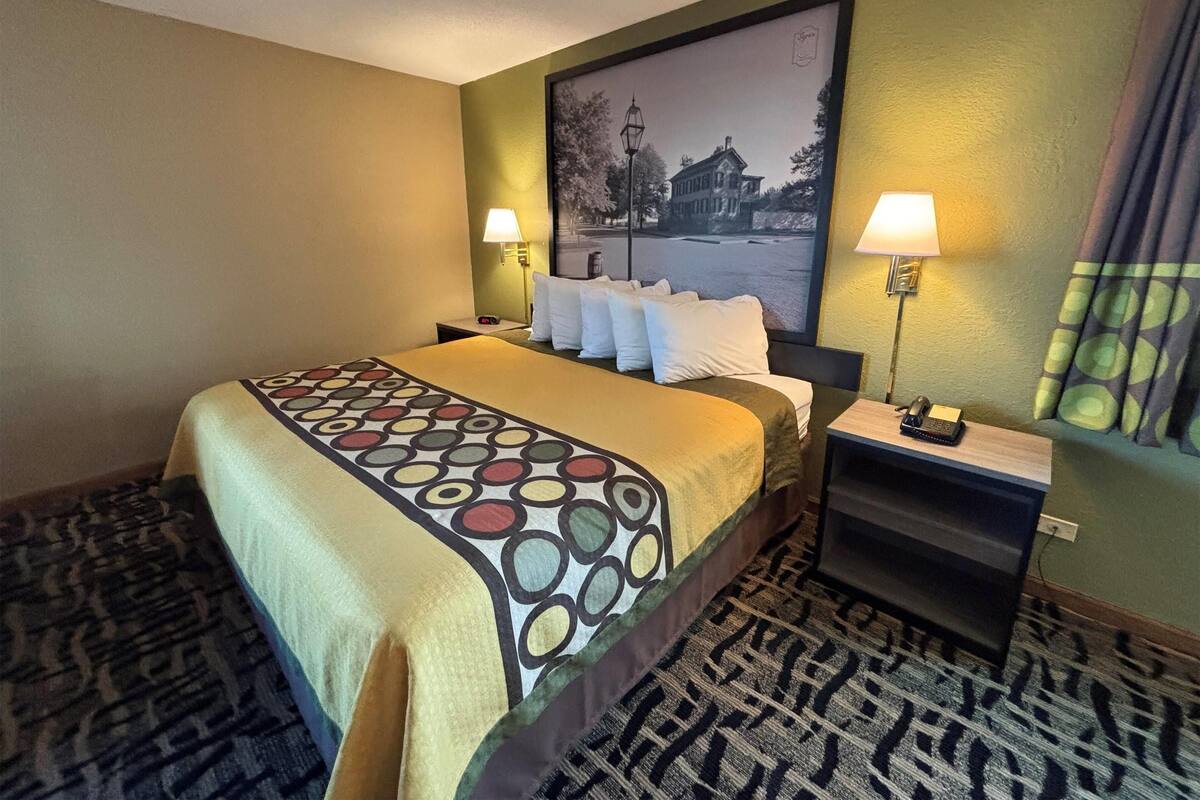 Ein Hotelzimmer mit einem Bett, zwei Nachttischen mit Lampen, einem gemusterten Teppich und einem großen, gerahmten Bild an der Wand.