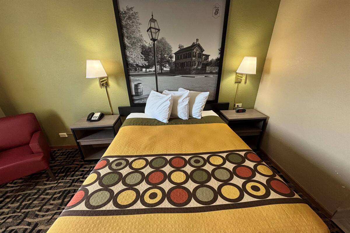 Ein Hotelzimmer mit einem Bett, zwei Nachttischlampen, einem Nachttisch mit Telefon, einem roten Sessel und einem gerahmten Bild an der Wand.