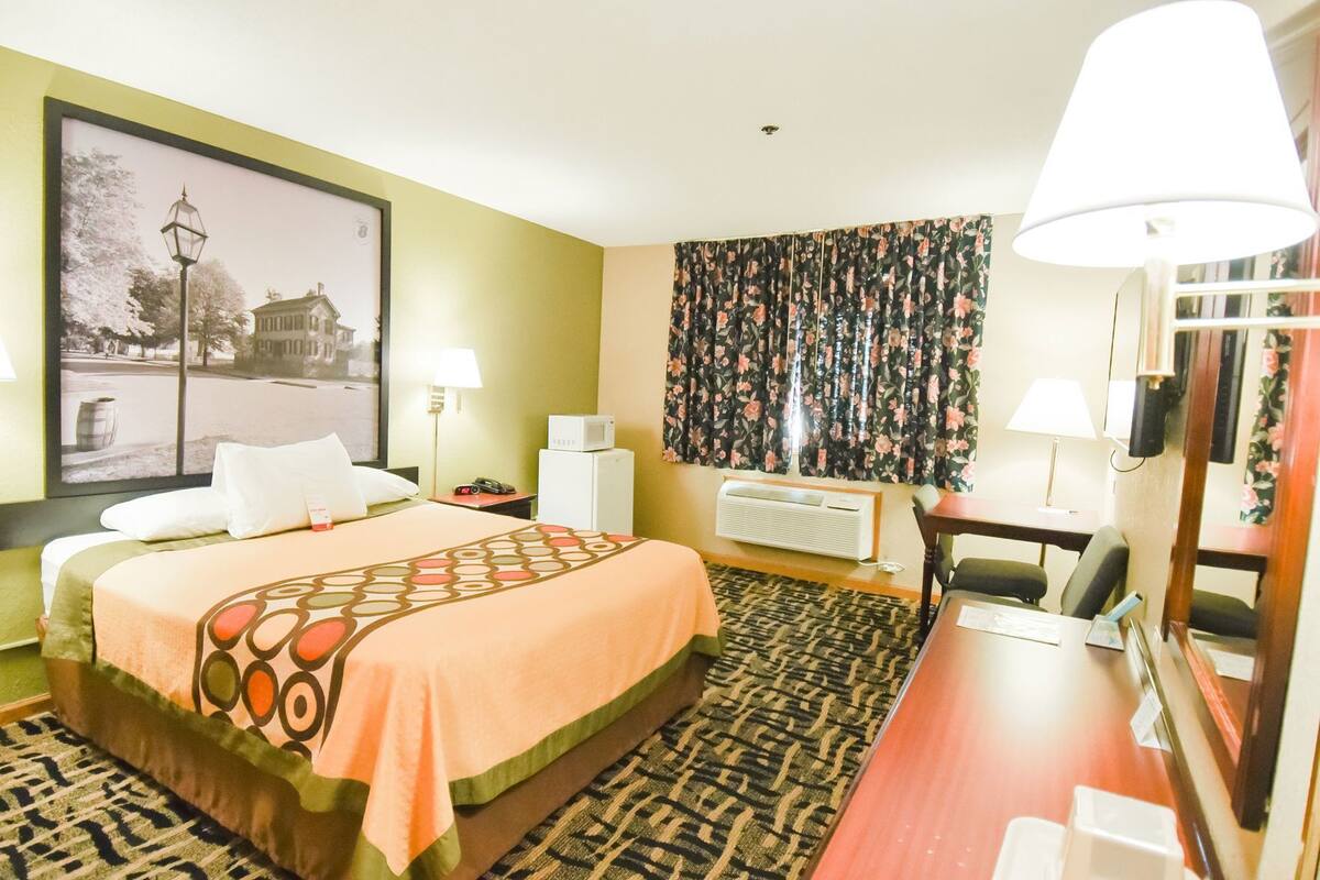 Ein Hotelzimmer mit Bett, Schreibtisch, Stuhl, Heizkörper, Lampe, einem Bild an der Wand und einem Fenster mit Vorhängen.
