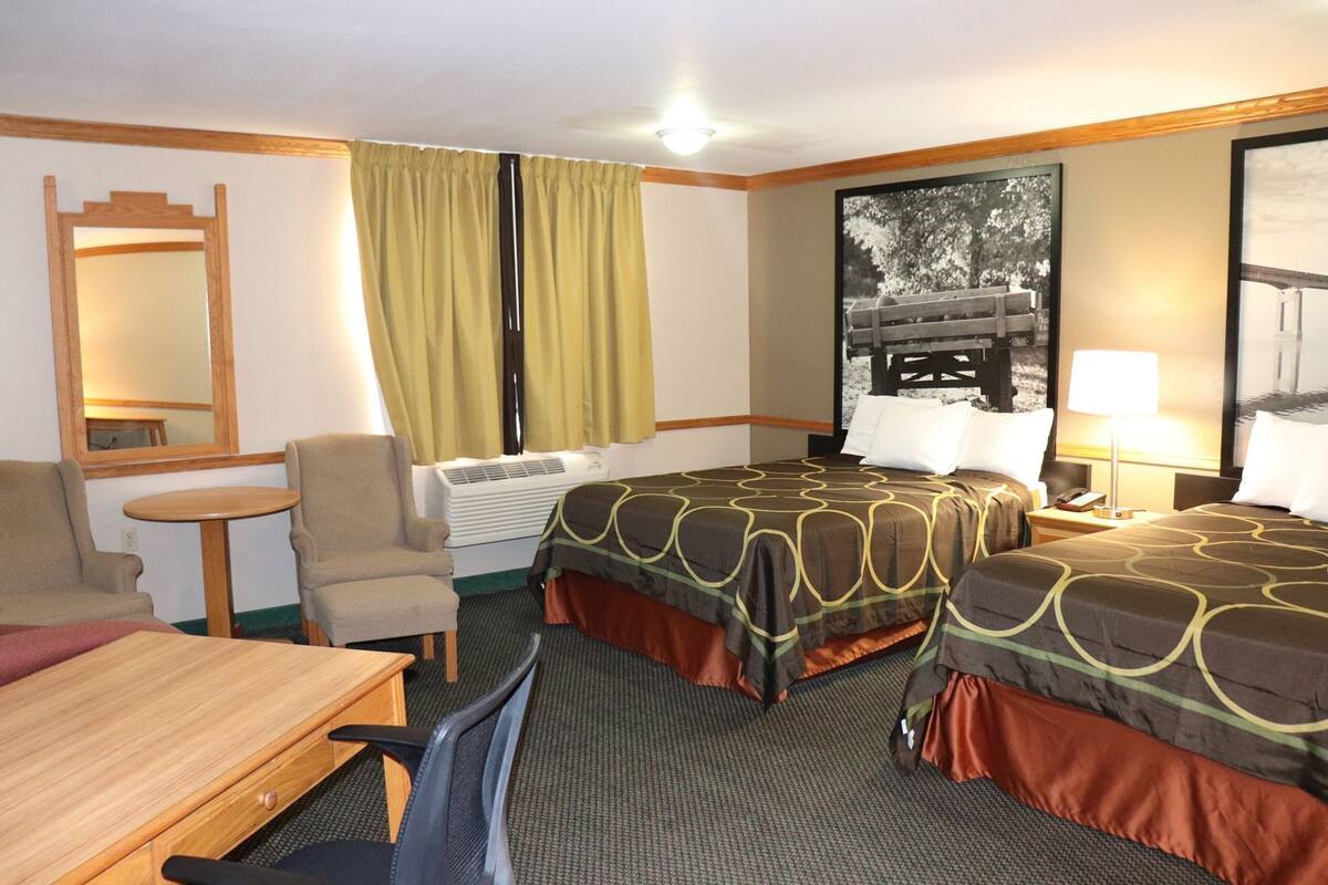 A hotel room with two beds, a desk, a chair, a small table, a mirror, a window with curtains, and a wall-mounted air conditioner.