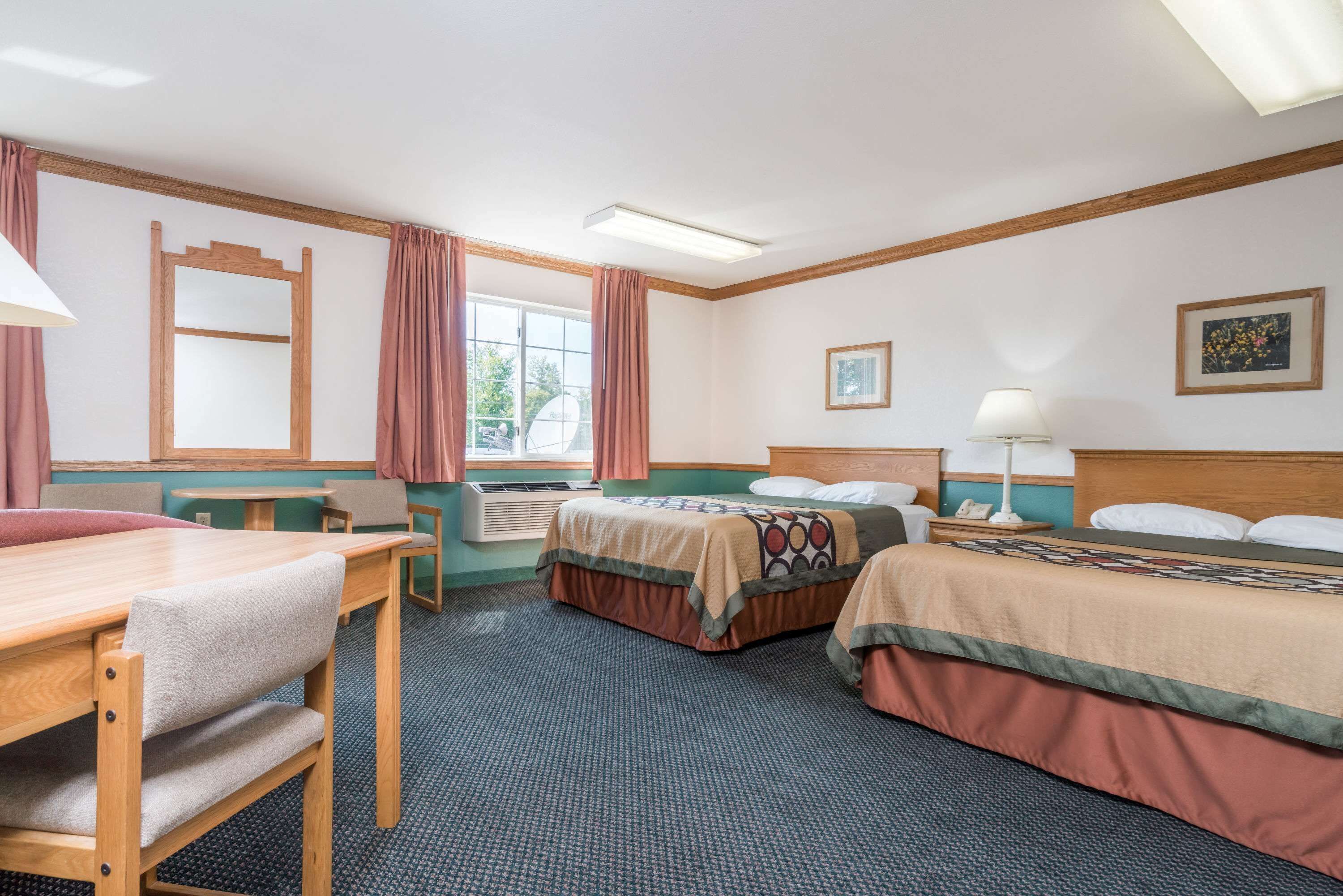 A hotel room with two beds, a dining table, chairs, a window with curtains, a ceiling fan, and a framed picture on the wall.
