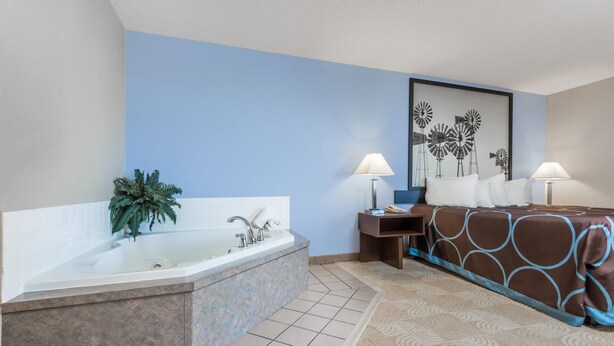 A hotel room with a bathtub, a bed with a patterned bedspread, a nightstand with a lamp, and a wall with a windmill painting. - Super 8 by Wyndham Beloit KS (Beloit)
