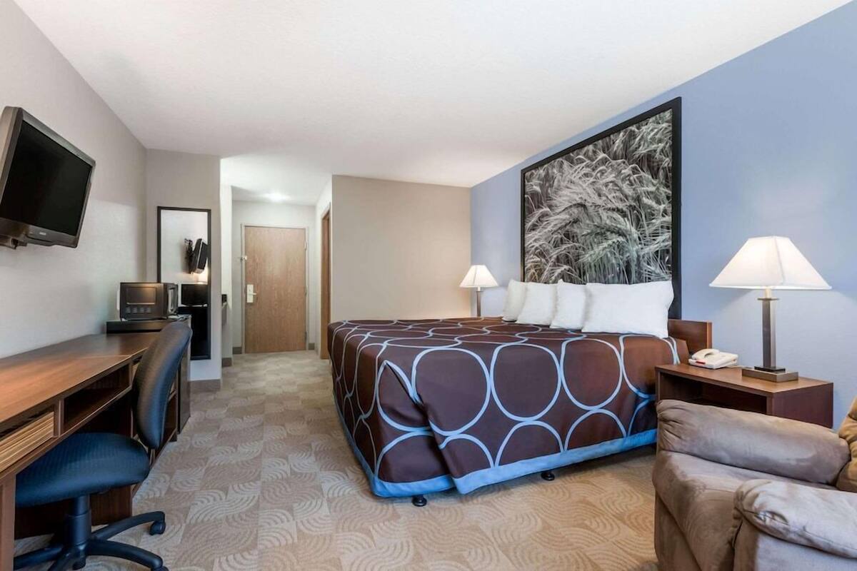 A hotel room with a bed, desk, chair, TV, and artwork on the wall.