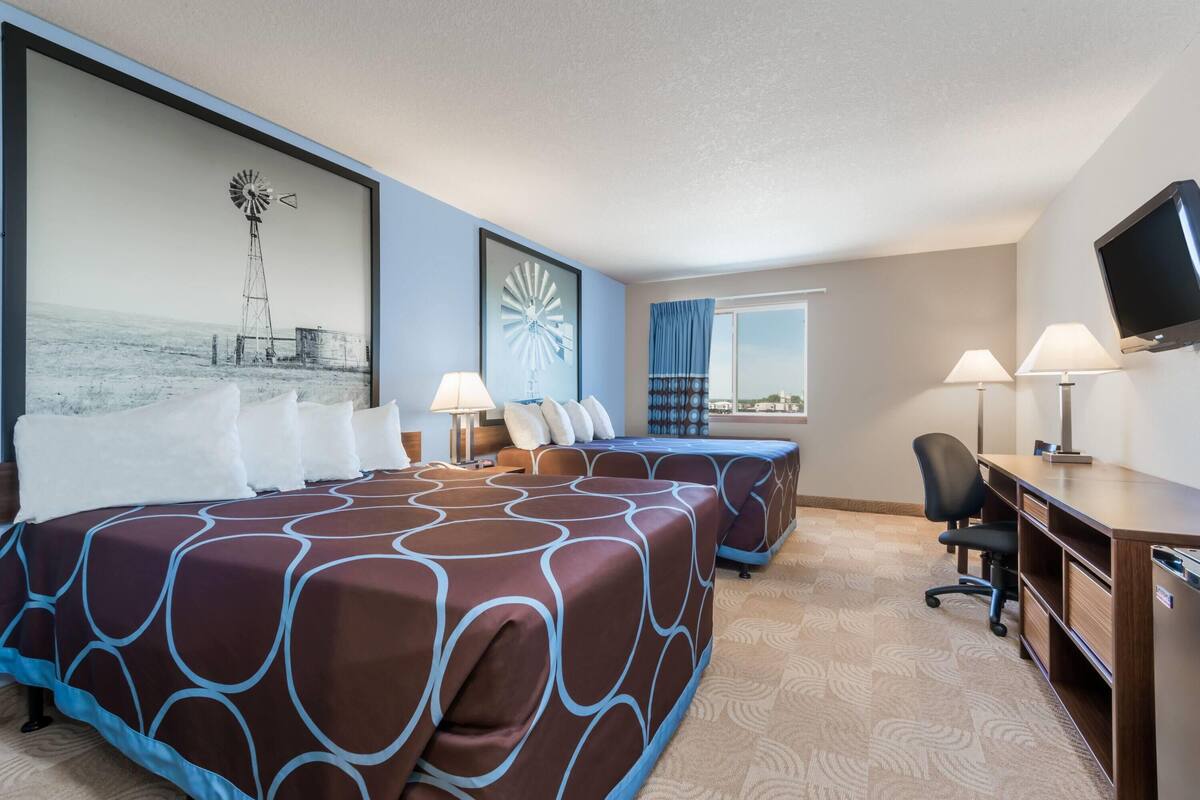 A hotel room with two beds, a desk, a chair, a TV, and a large window with curtains.