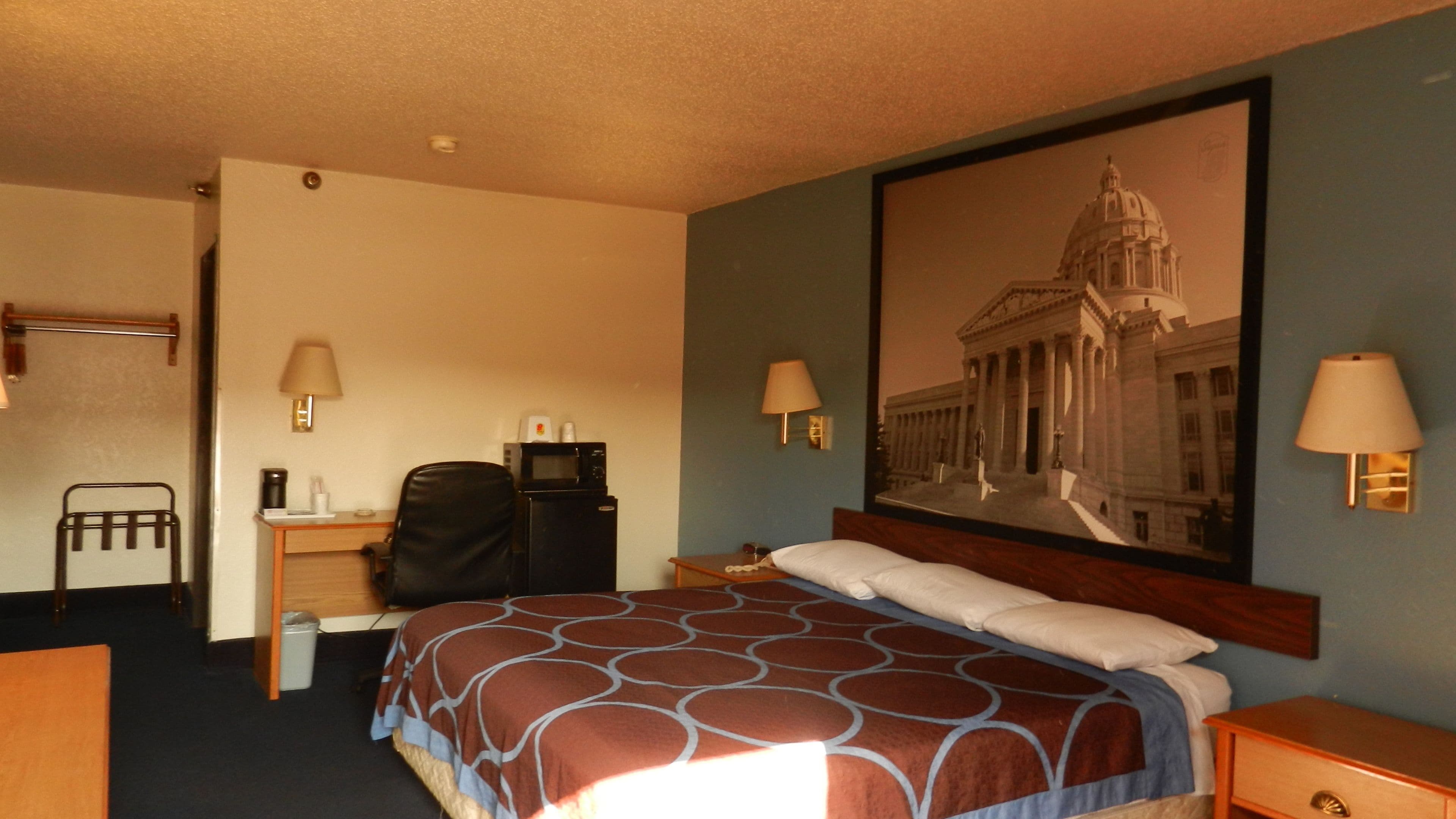 A hotel room with a bed, a desk, a chair, a lamp, and a large framed picture of a building.