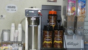 Una zona de desayuno con una cafetera, dos dispensadores de café con la etiqueta 'Farm Brothers' y cajas de cereales que incluyen 'Raisin Bran' y 'Frosted Flakes'.