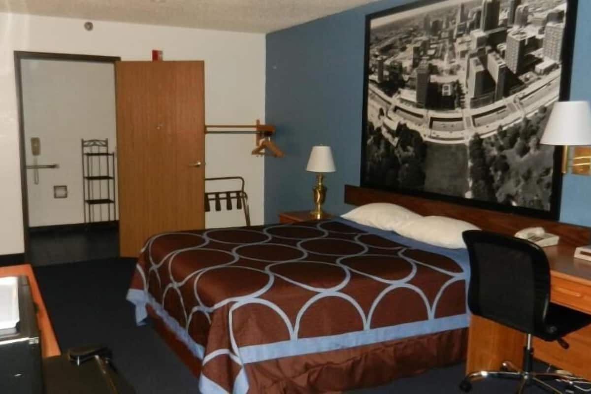 A hotel room with a bed, desk, chair, lamps, and a large cityscape painting on the wall.