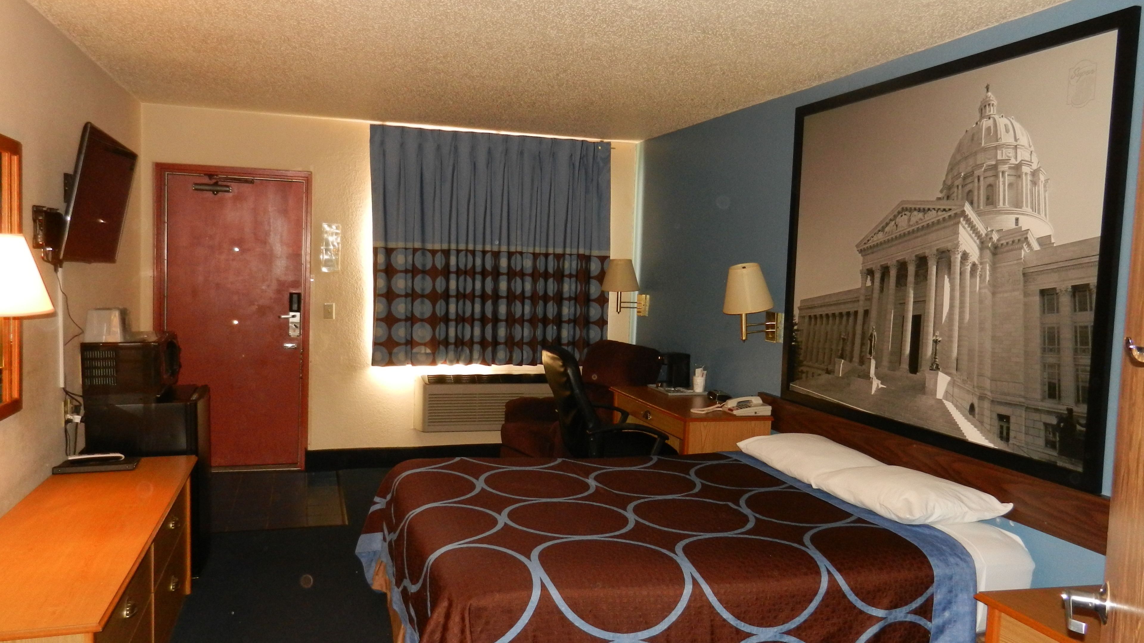A hotel room with a bed, a desk, a chair, a TV, and a large framed picture on the wall.
