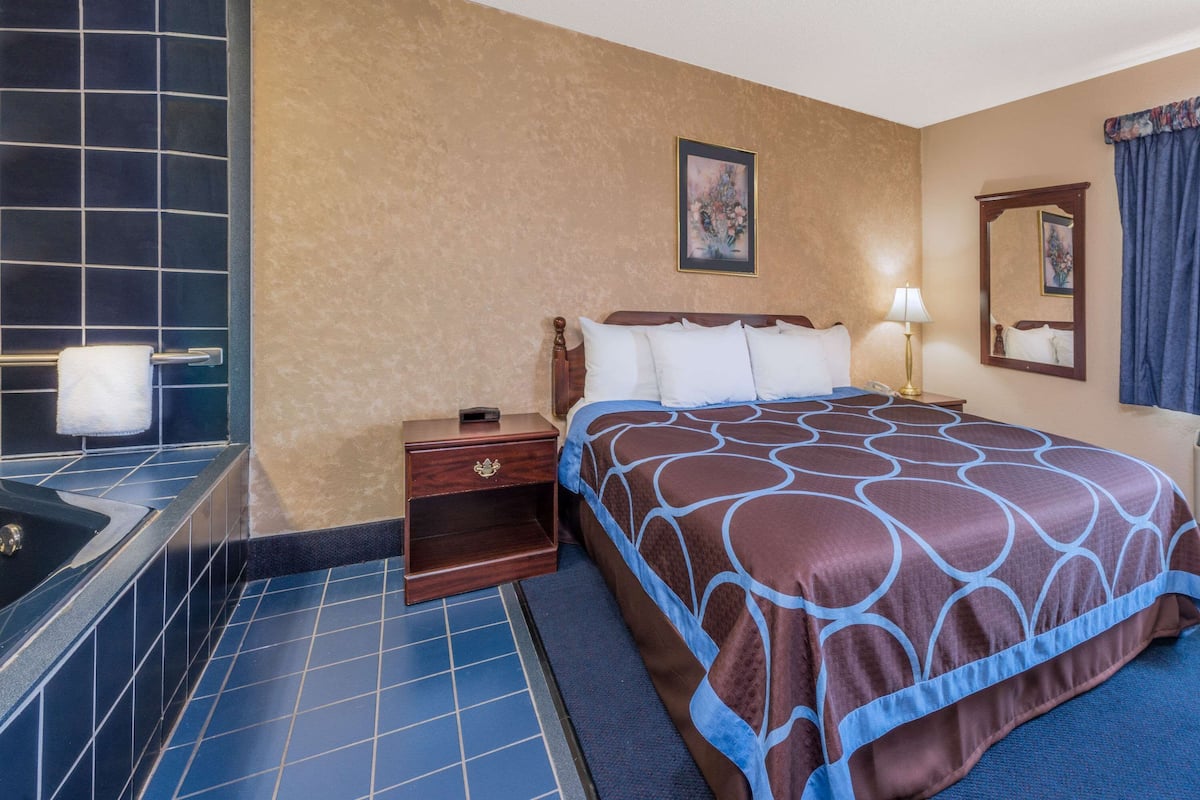 A hotel room with a large bed, a bedside table, a nightstand, a bathroom with a bathtub, and a mirror on the wall.