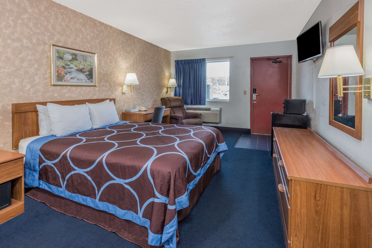 A hotel room with a large bed, a desk with a lamp, a chair, a TV, and a door.