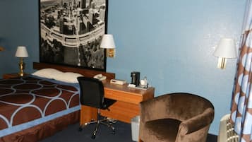 A hotel room with a bed, a desk, a chair, a coffee maker, and a large cityscape painting on the wall.