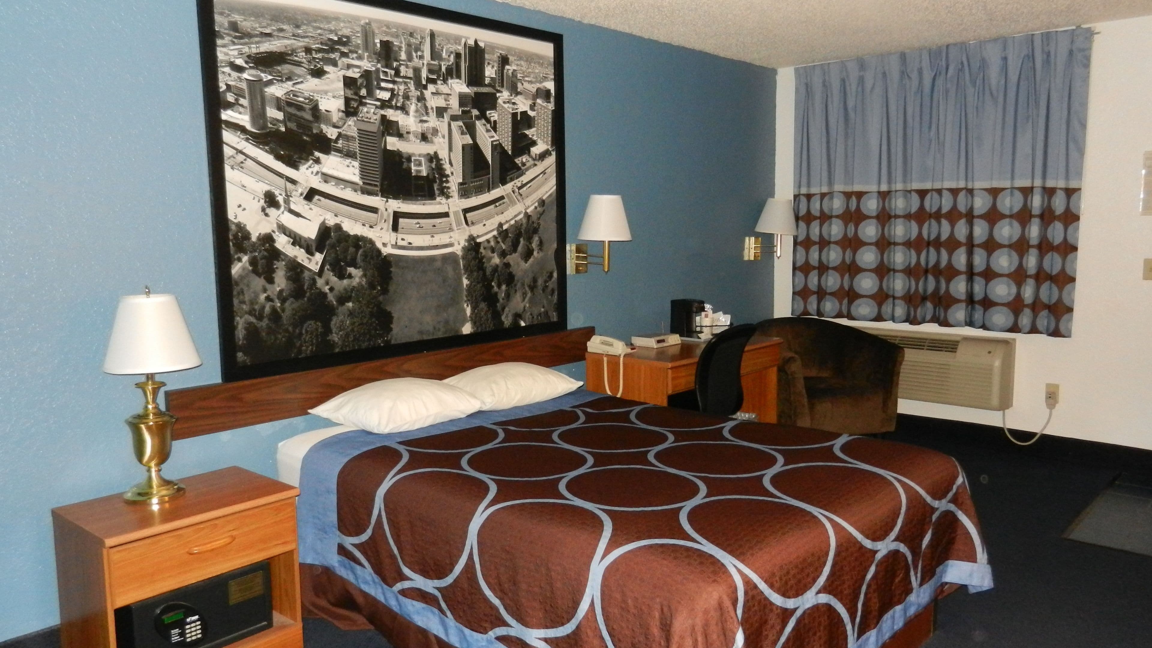 A hotel room with a bed, two bedside lamps, a desk with a telephone, a wall-mounted air conditioner, and a large cityscape painting.