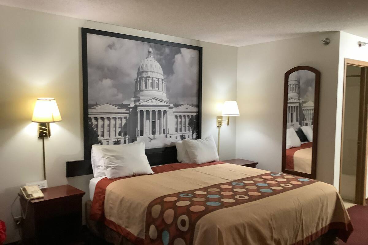 A hotel room with a large bed, two bedside lamps, a desk with a phone, and a large framed picture of a building on the wall.