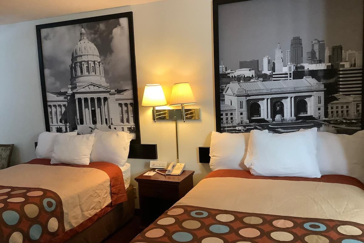 A hotel room with two beds, a nightstand with a lamp, and two large black and white cityscape posters on the walls.