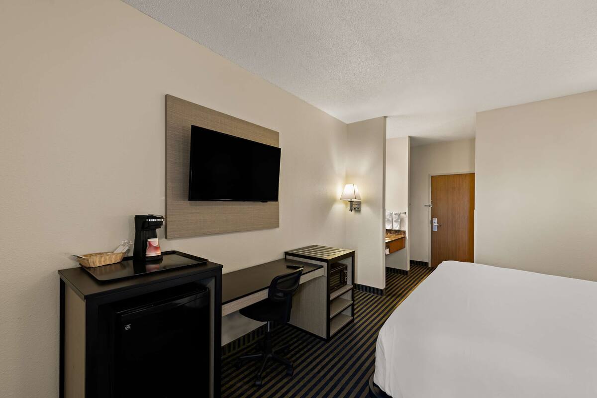 A hotel room with a bed, a desk with a chair, a television mounted on the wall, a coffee machine, and a small refrigerator.