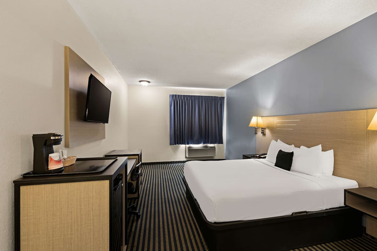 A hotel room with a bed, a desk with a coffee maker, a TV mounted on the wall, and a window with curtains.