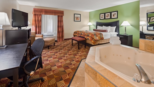 A hotel room with a large bed, a desk with a computer, a TV, a bathroom with a bathtub, and a patterned carpet.