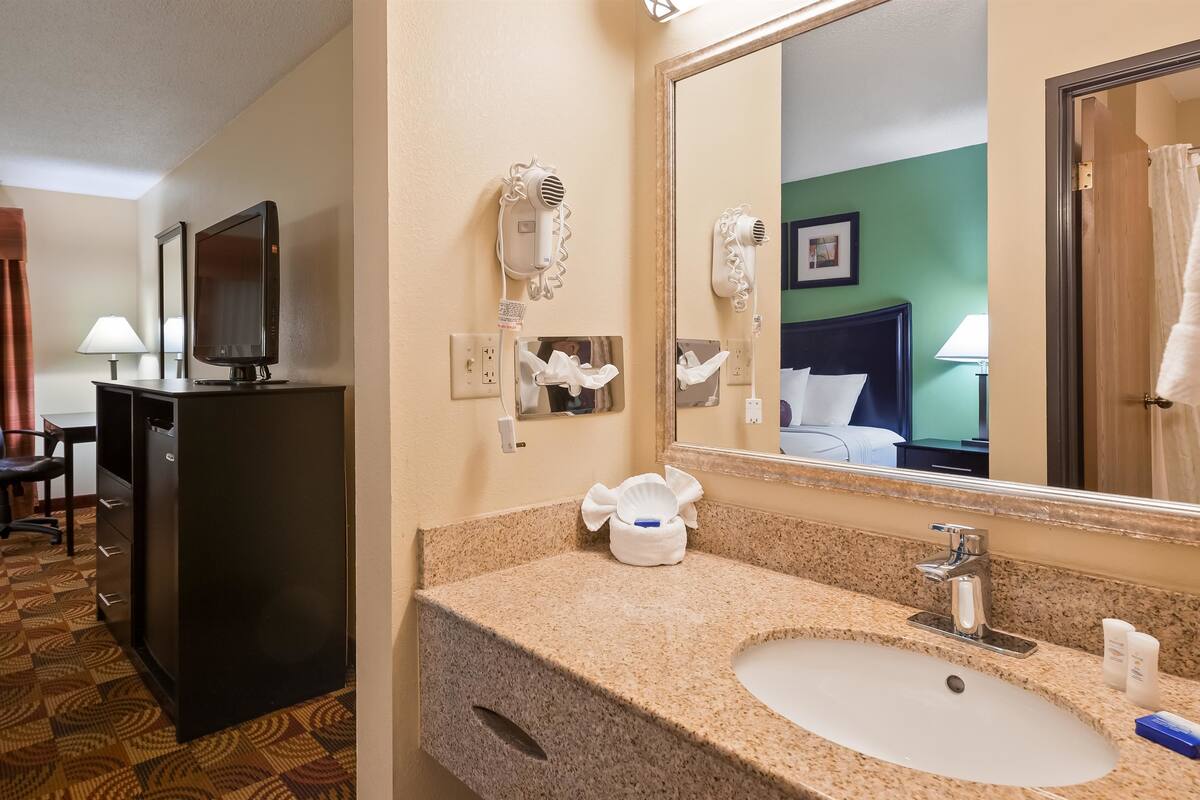 Hotelkamer met een bed, een bureau met stoel, een televisie, een haardroger, een spiegel en een badkamer met wastafel en toiletartikelen.