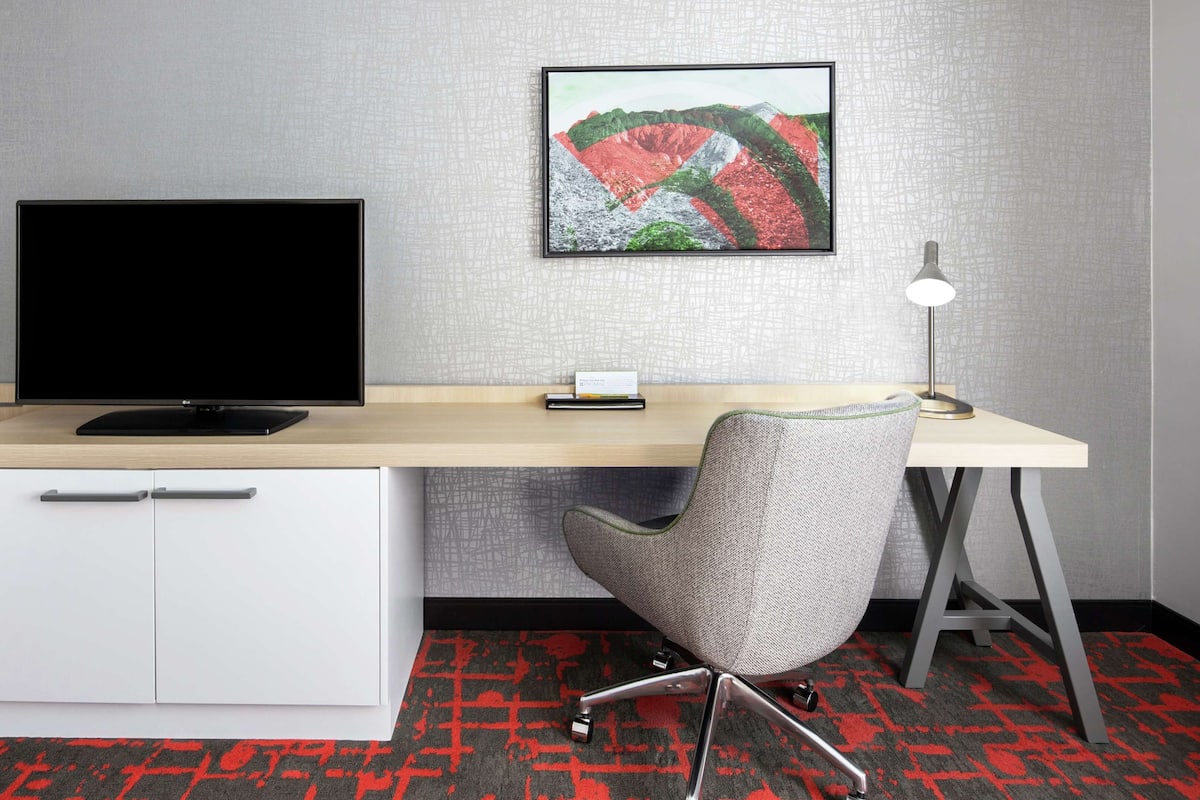 Habitación de hotel con escritorio, televisión, silla, lámpara de escritorio y alfombra.
