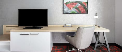 A hotel room with a desk, a television, a chair, a desk lamp, and a carpet.