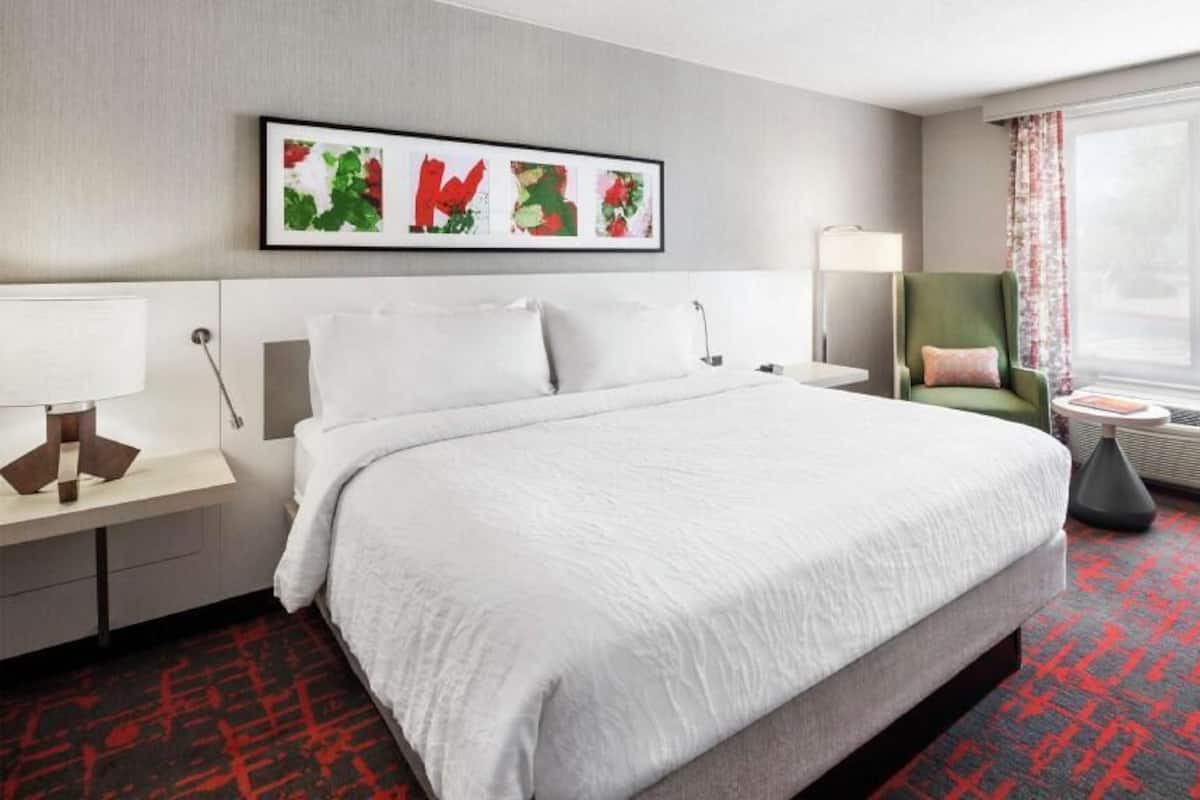 Una habitación de hotel con una cama grande, una mesita de noche, una silla, una ventana con cortinas y una obra de arte colgada en la pared.