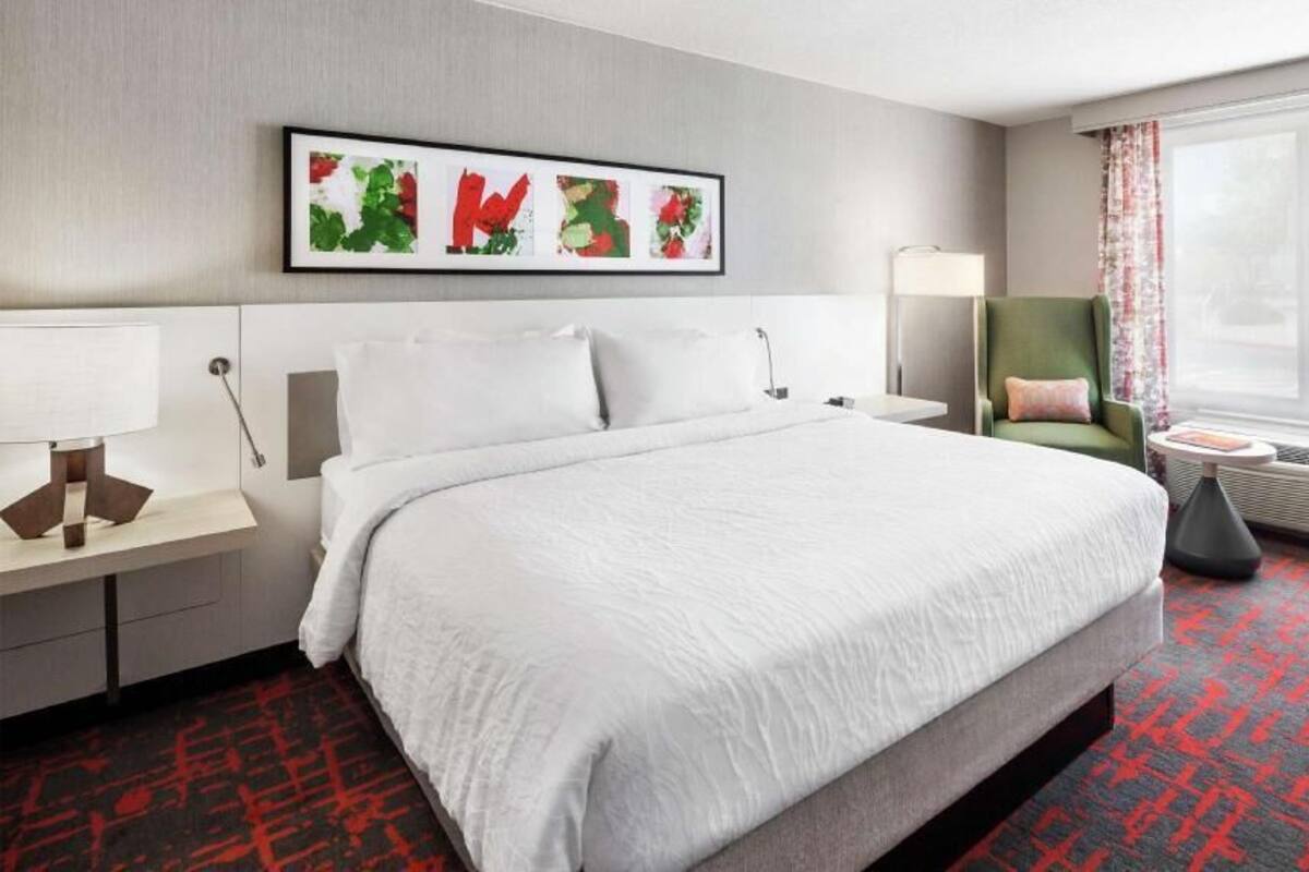 Habitación de hotel con una cama grande, un escritorio con lámpara, una silla, un ventanal con cortinas y una obra de arte colgada en la pared.