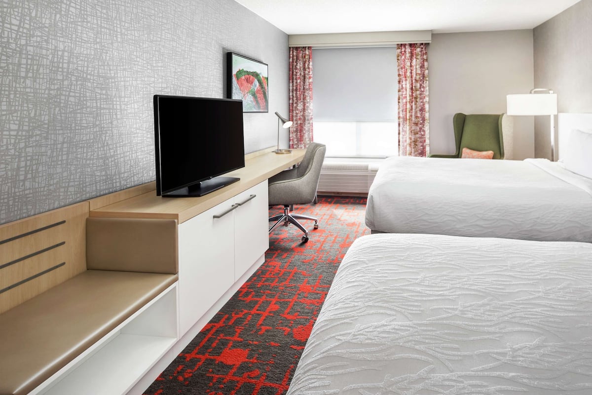 Habitación de hotel con televisor de pantalla plana, escritorio, dos camas, una silla y ventana con cortinas.
