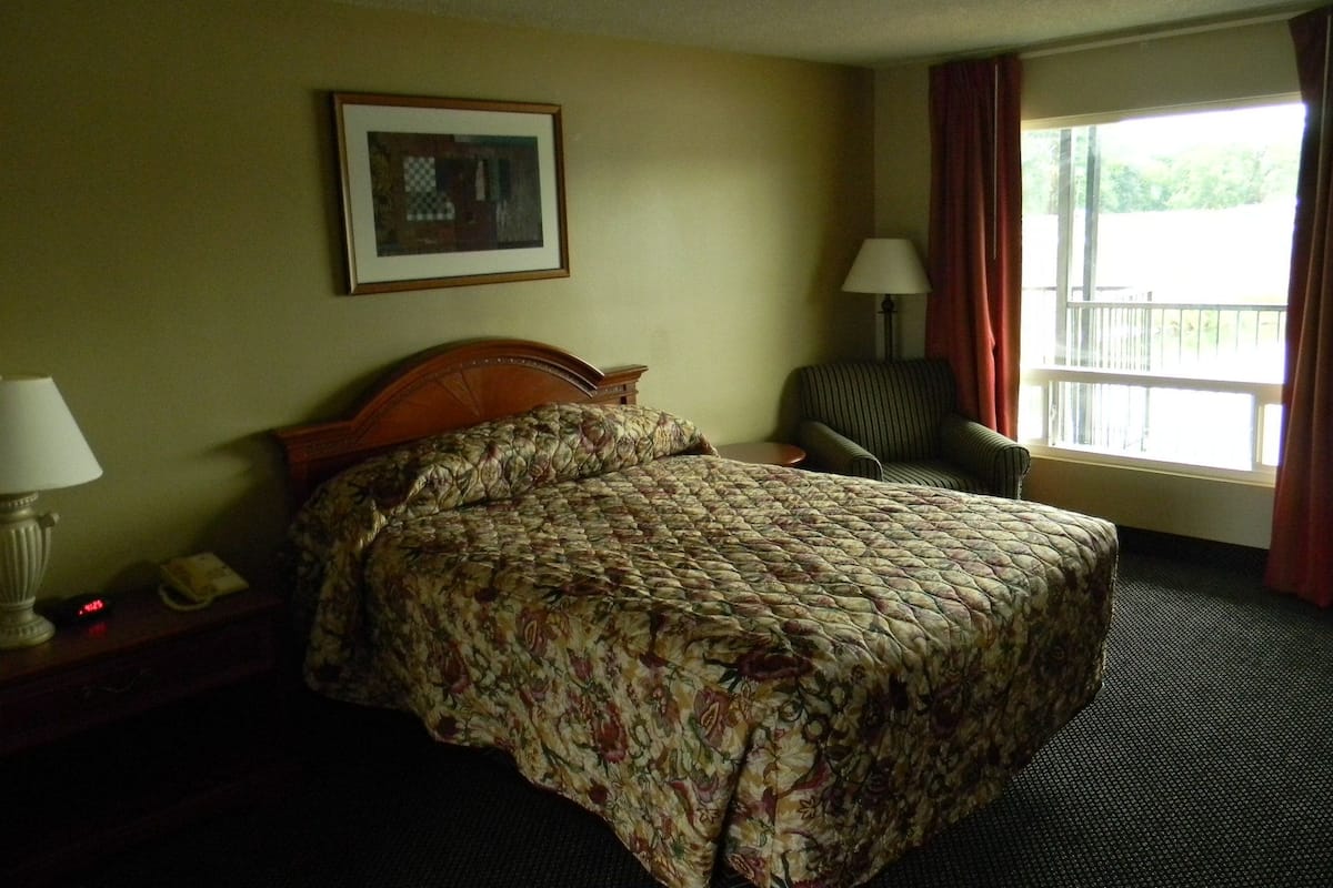 A hotel room with a bed, a chair, a nightstand, a lamp, a painting on the wall, and a window with curtains.