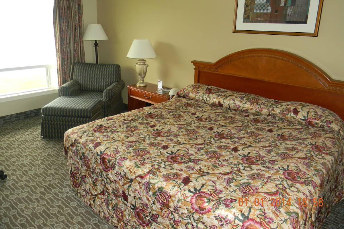 A hotel room with a large floral bedspread, a wooden headboard, a chair, a lamp, and a framed picture on the wall.