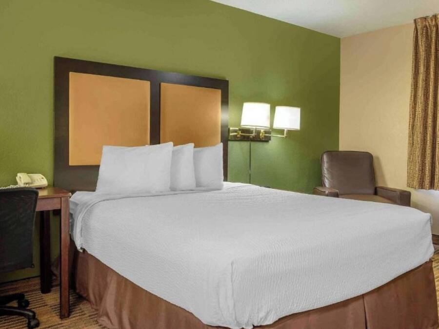 A hotel room with a large bed, a desk with a telephone, a chair, and a wall-mounted lamp.