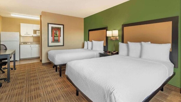 A hotel room with two beds, a desk, a kitchenette, and a framed picture on the wall.