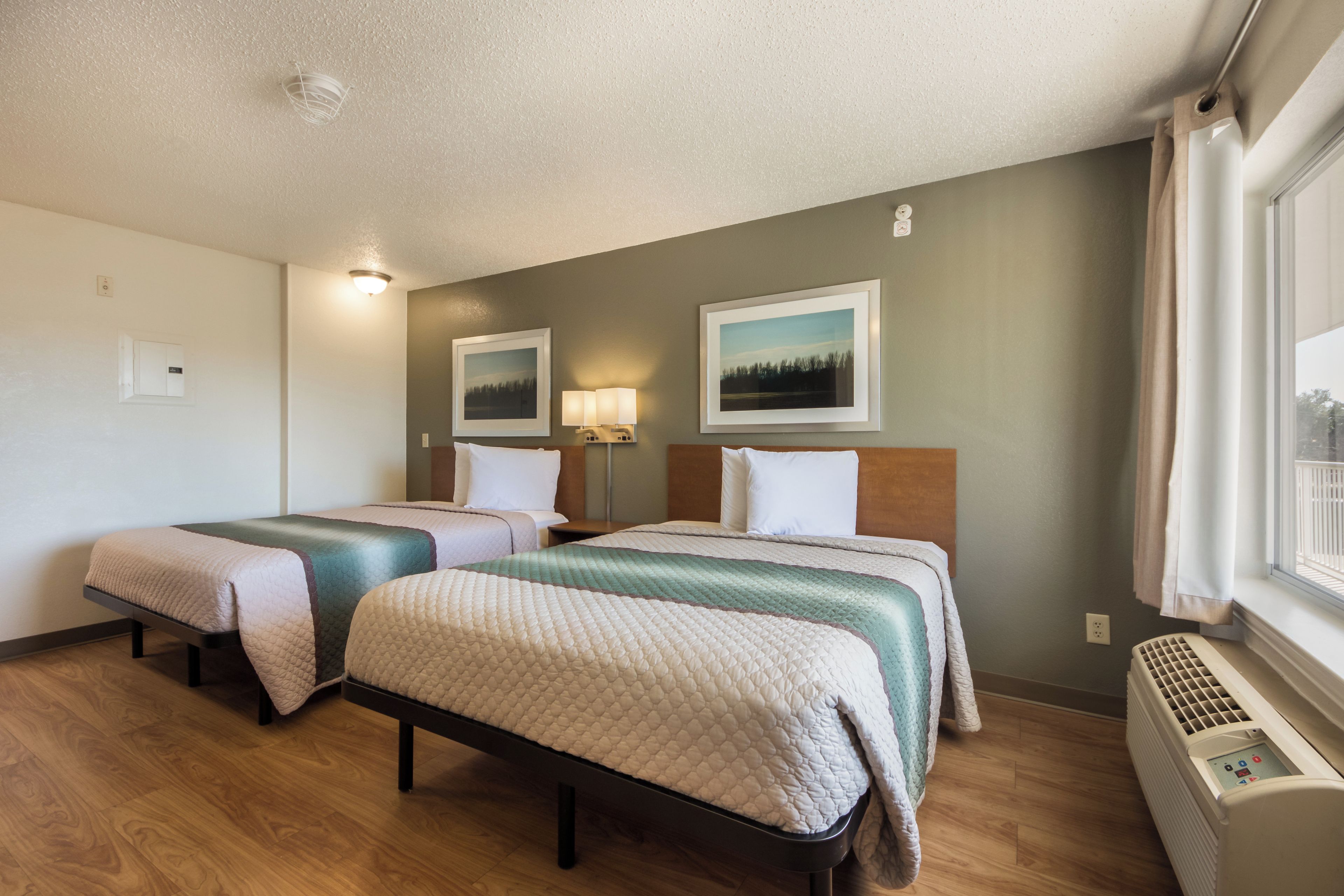 Habitación de hotel con dos camas, una ventana con cortinas, un calentador fijado a la pared y dos cuadros enmarcados.