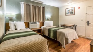 Een hotelkamer met twee bedden, een bureau, een stoel, een tafel, een douchegordijn, een wandairconditioner, een wandlamp en een lijst met een foto aan de muur.