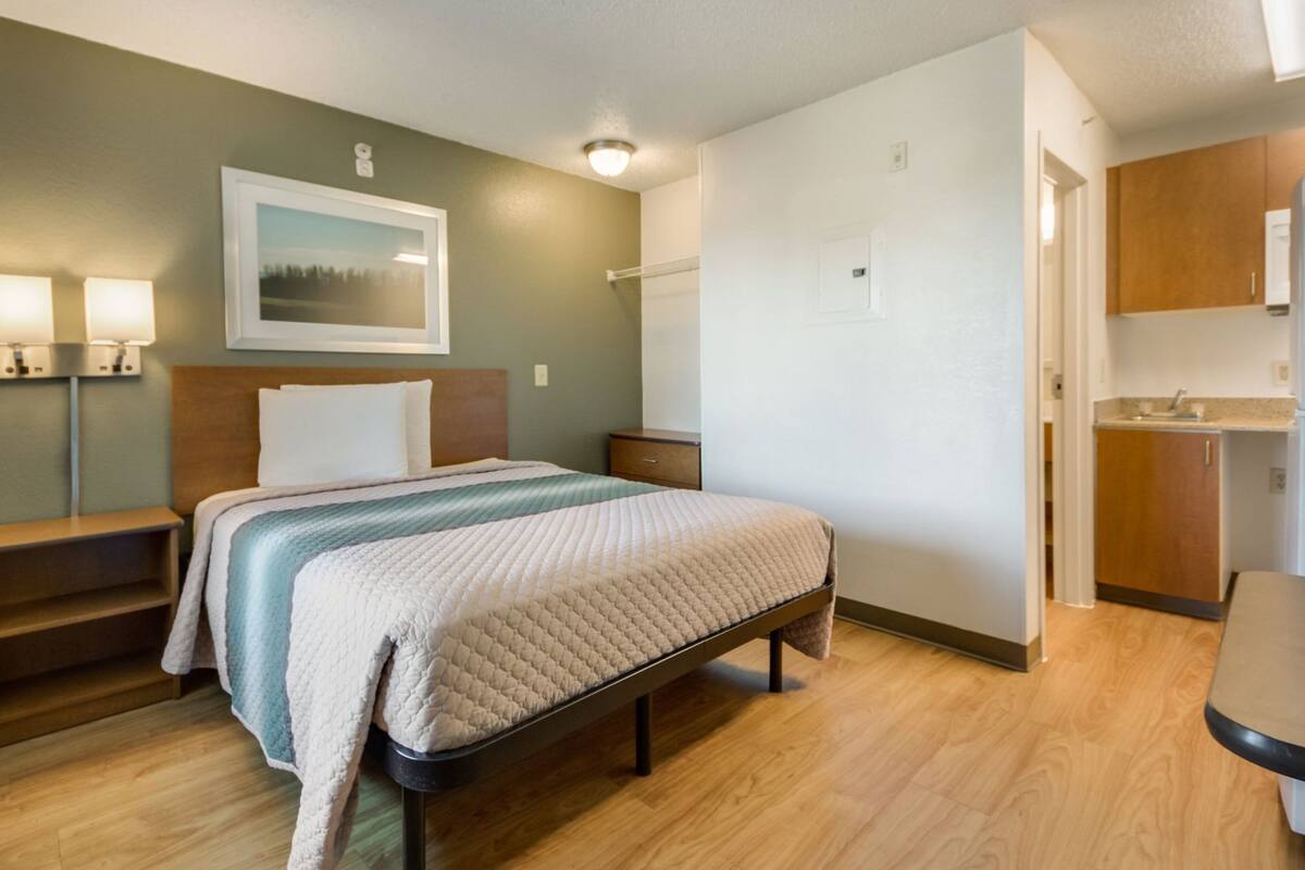 Een slaapkamer met een bed, nachtkastjes, een klein kastje, een foto aan de wand en een kitchenette.