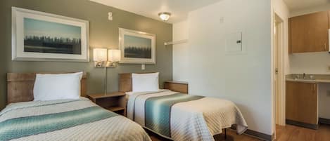 Una habitación de hotel con dos camas, una mesita de noche, una lámpara, un cuadro, un armario y una kitchenette.