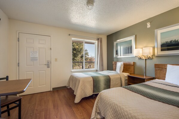 A hotel room with two beds, a wooden table, a window with a view, and a painting on the wall.
