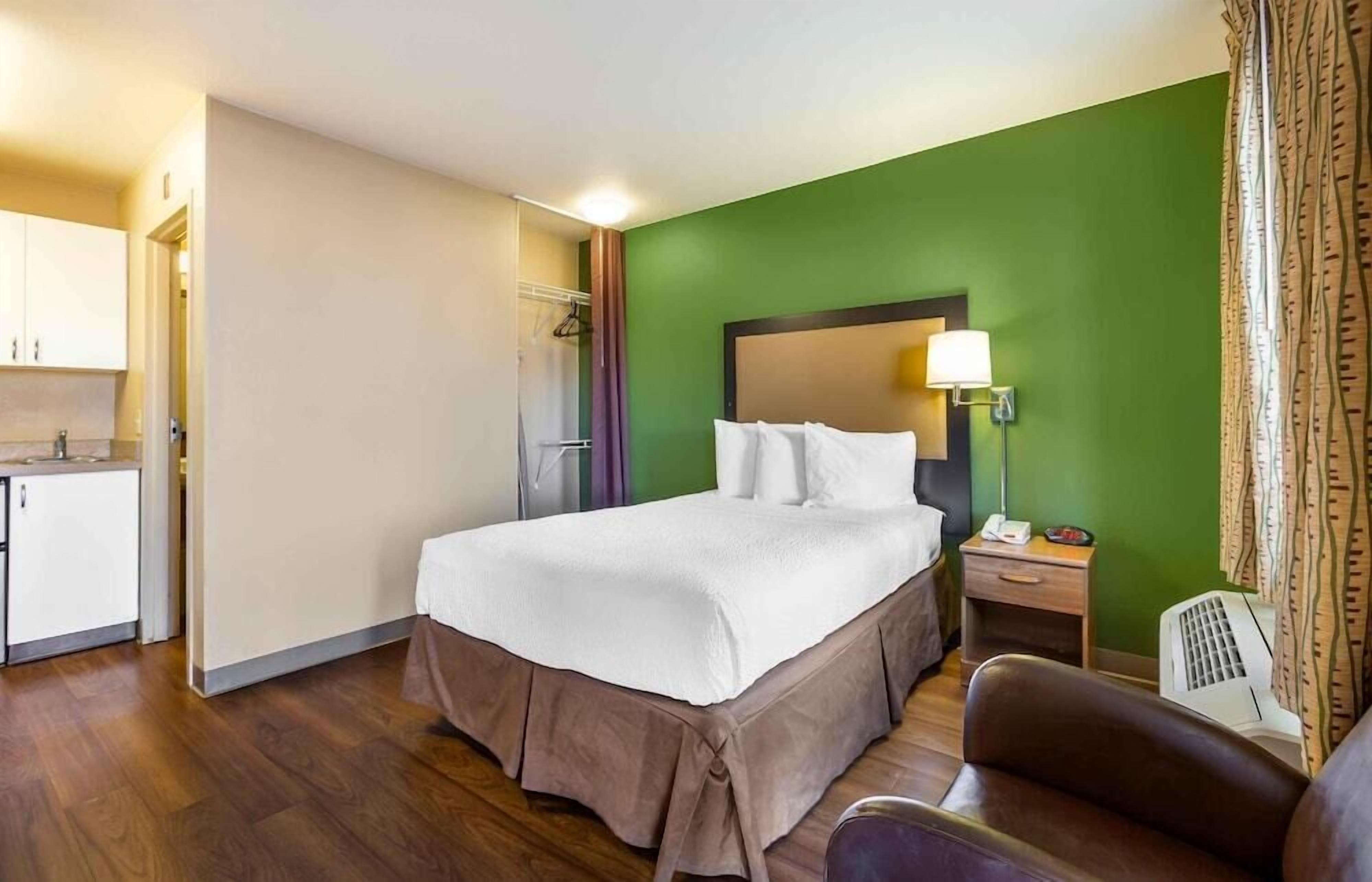 A hotel room with a bed, bedside table, lamp, chair, and a kitchenette area.