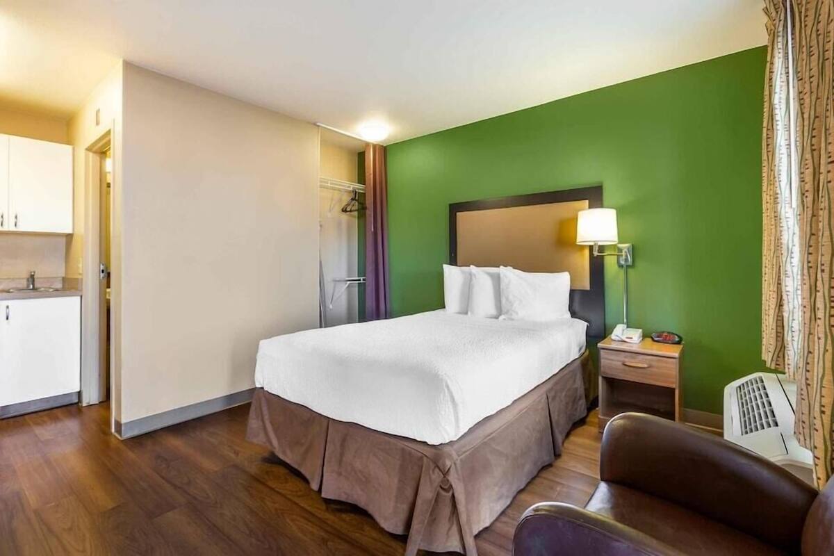 A hotel room with a bed, bedside table, lamp, chair, and a kitchenette area.
