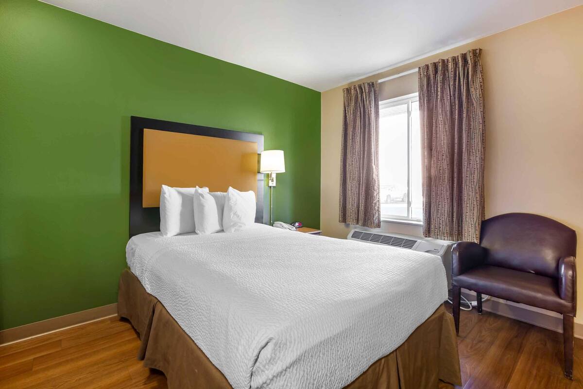 A hotel room with a large bed, a desk with a lamp, a chair, and a window with curtains.