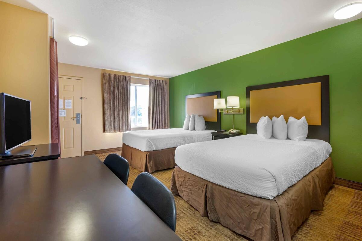 A hotel room with two beds, a TV, a desk, and a window with curtains.